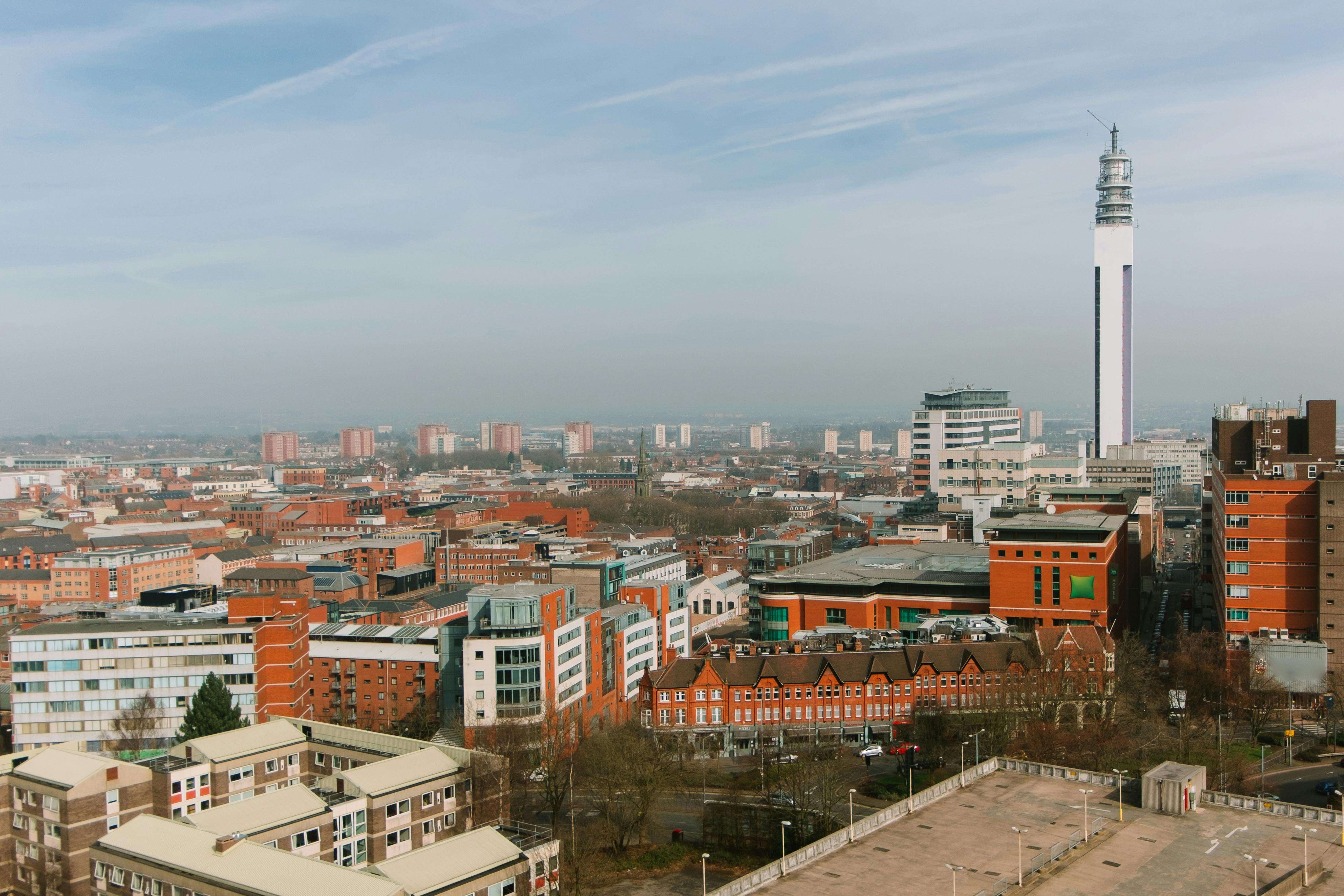 A panoramic urban vista unfolds under a soft, hazy sky streaked with delicate white clouds, casting a muted, diffused light over the cityscape. Dominating the skyline on the right is the towering, slender black-and-white structure of the BT Tower, its metallic sheen catching the ambient light, standing as a stark vertical contrast to the dense, low-to-mid-rise architecture below. The city is a tapestry of brick-red and terracotta-colored buildings, many with traditional gabled roofs and rows of windows, interspersed with modern glass-and-steel edifices that gleam faintly. Streets and avenues weave through clusters of residential and commercial structures, their surfaces mostly subdued in earthy tones—ochres, creams, and grays—with occasional bright accents like green glass panels or a red car parked near a building. Bare-branched trees suggest a winter or early spring setting, and a large, open paved plaza in the foreground, bordered by low railings and lampposts, remains empty, emphasizing the urban expanse. The overall mood is calm and slightly nostalgic, as if captured from a high vantage point overlooking a city that blends its historic character with modern ambition.